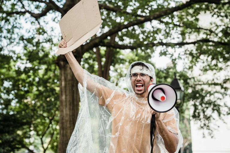 Man holding megaphone and cardboard at protest rally in park.